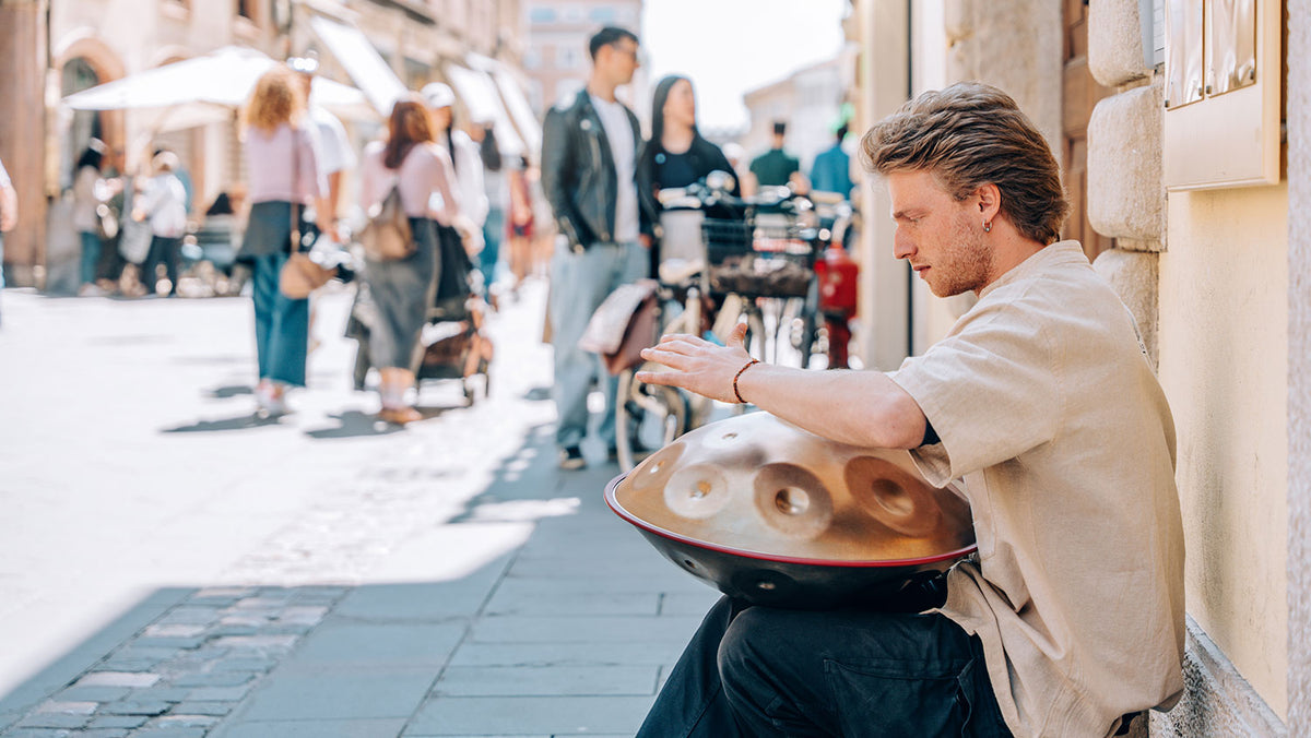 A young man sits against a house wall on a busy shopping street and plays a handpan on his lap.
